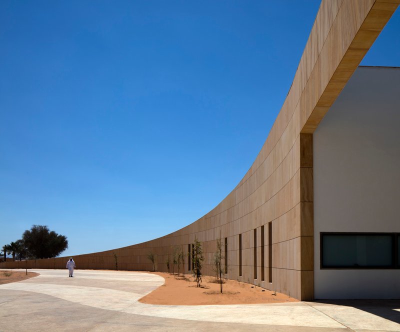 Curved sandstone wall with vertical window openings and a figure walking along the pathway