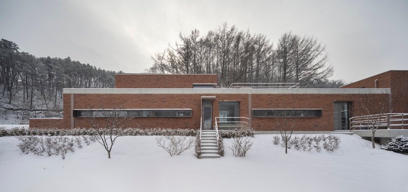 Horizontal brick facade with ribbon windows and front stair in a snow-covered garden beneath winter trees