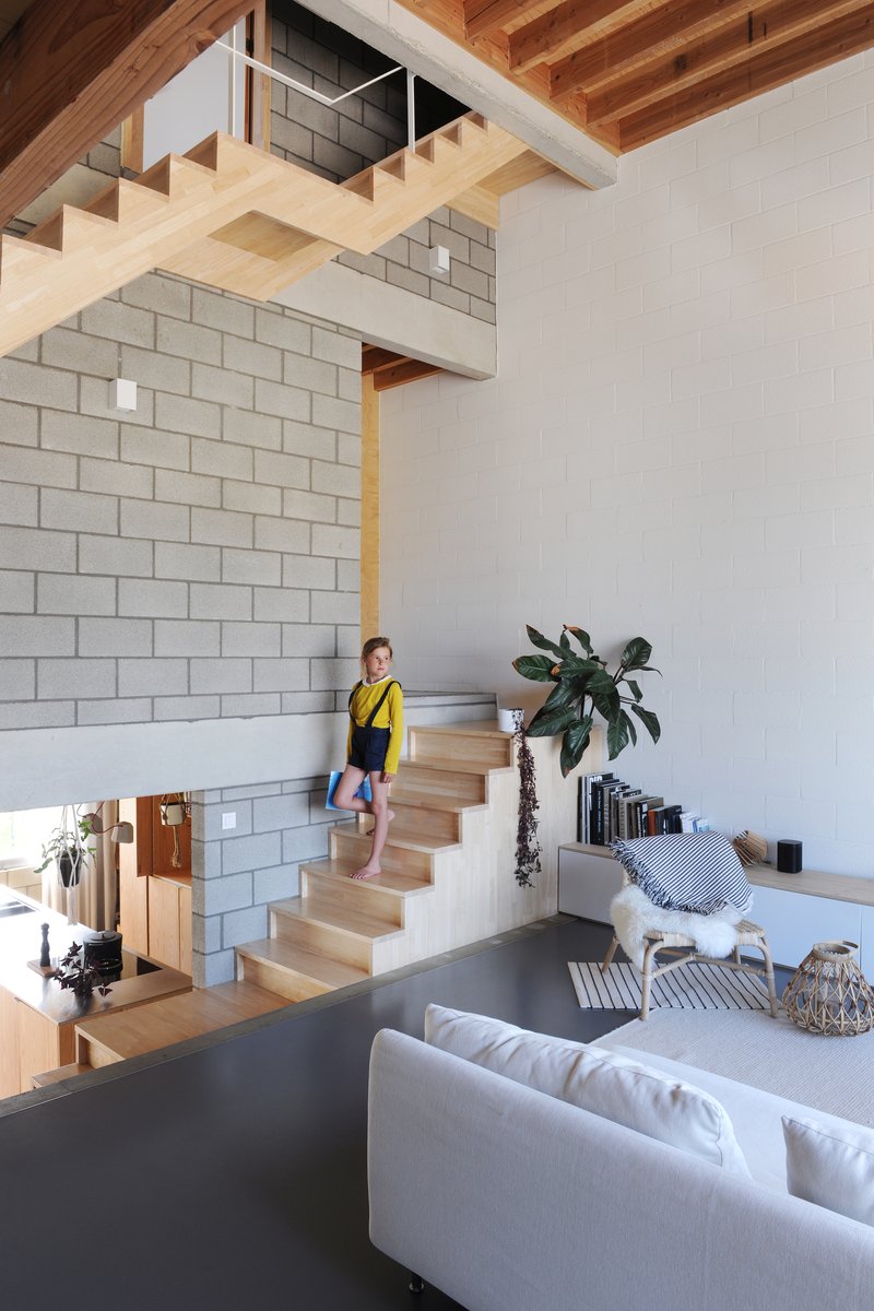 Double-height living space with open timber staircase and child ascending beside concrete block wall
