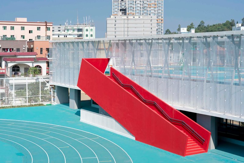 Red sculptural staircase with angled treads rising above the turquoise running track and translucent facade panels