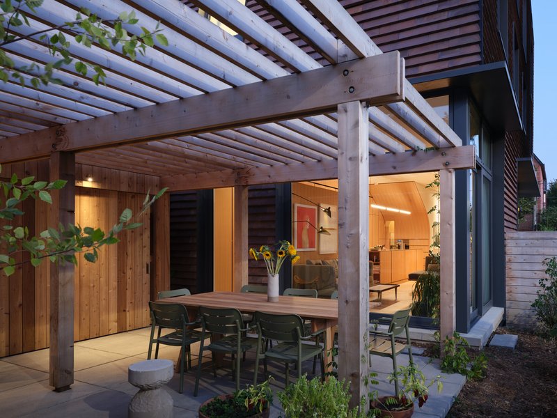 Outdoor dining area under timber trellis structure with potted plants and warm interior lighting