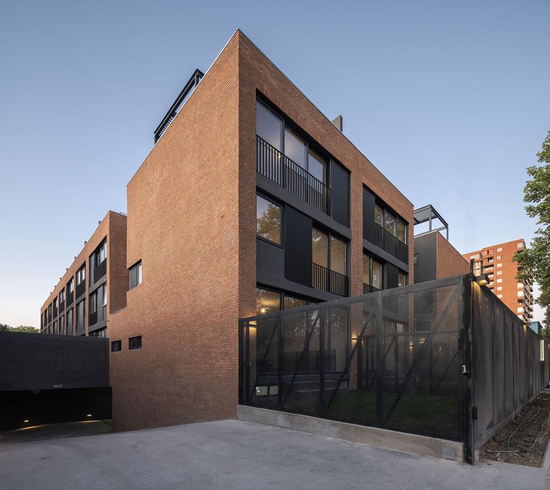 Corner elevation showing the brick volume with recessed balconies and translucent metal fence at dusk