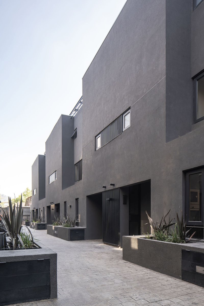Gray stucco facades with punched windows along a paved courtyard with planted concrete beds and succulents