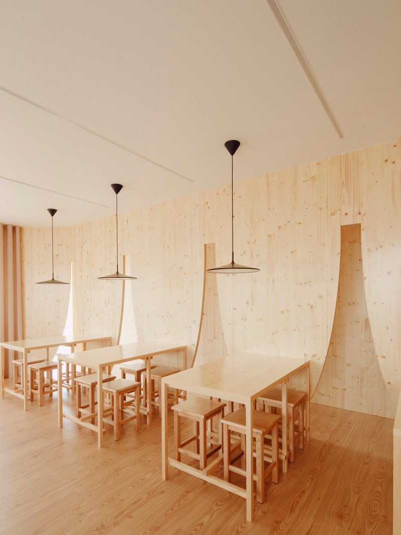 Communal dining tables with pendant lights and angled plywood wall panels beneath a white ceiling