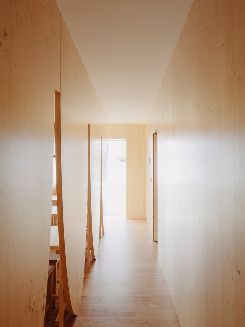 Narrow hallway with pale plywood walls and angled timber partitions opening to a sunlit doorway