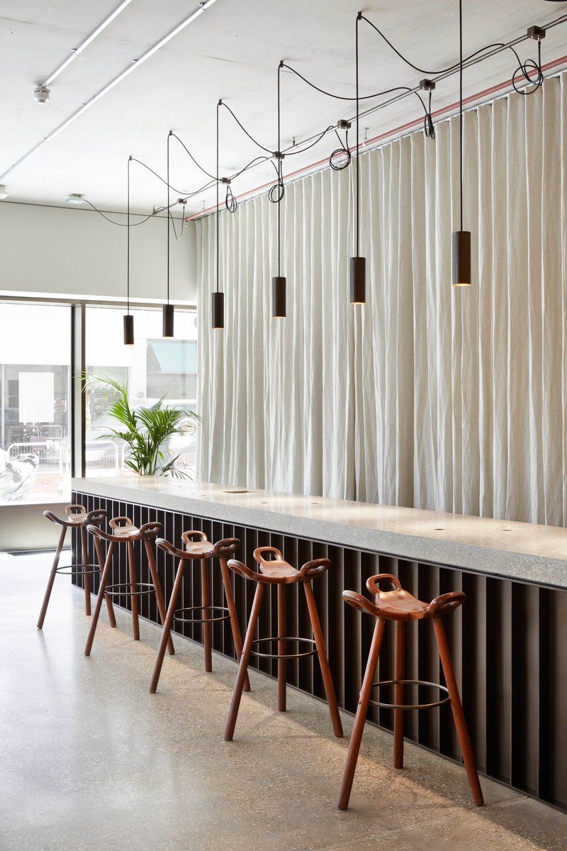 Long bar counter with timber stools below cylindrical pendant lights suspended from exposed conduit