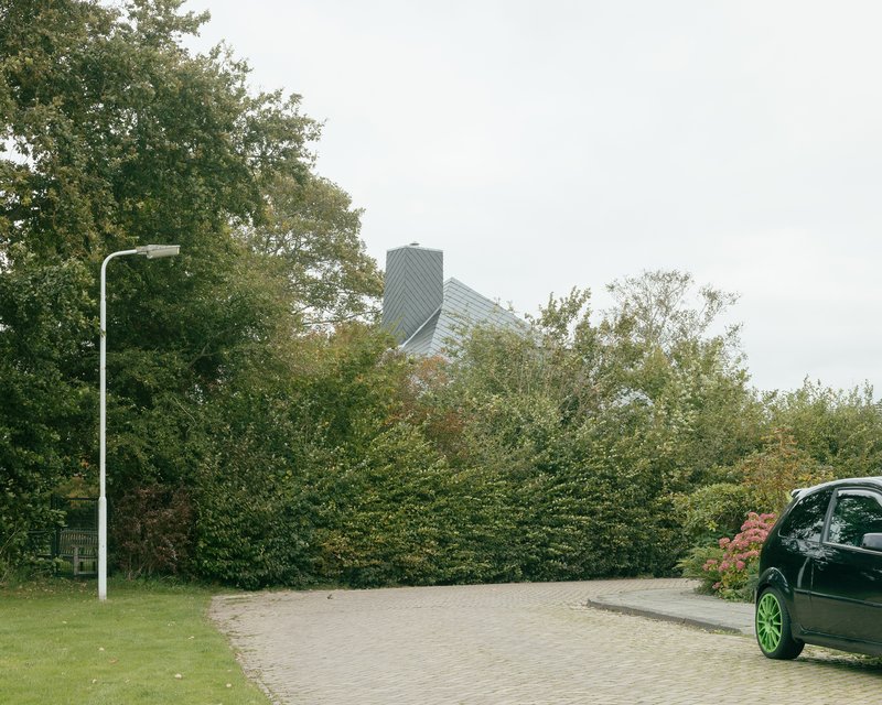 Street view showing the diamond-tiled roof rising above dense hedge and parked car
