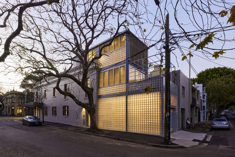 Oblique view from the street showing the illuminated glass brick volumes beside bare winter trees at twilight
