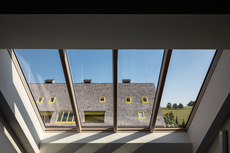 View through angled roof windows framing the grey shingle roof and yellow-trimmed dormers of the neighboring volume