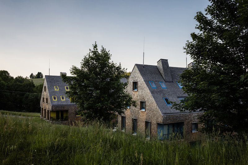 Two gabled volumes with slate roofs and stone cladding set in tall grass at dusk