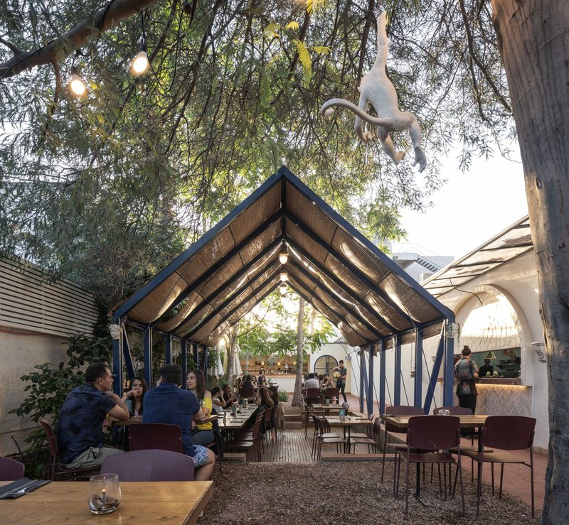 Outdoor dining courtyard with pitched timber canopy roof beneath overhanging tree branches at dusk