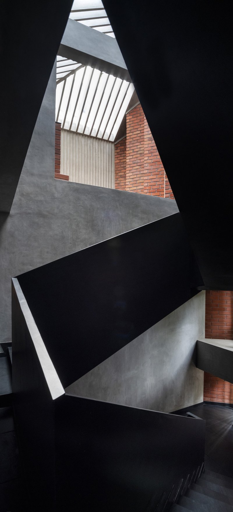 Vertical view up the stairwell showing triangular concrete landings and black steel flights beneath slatted skylight