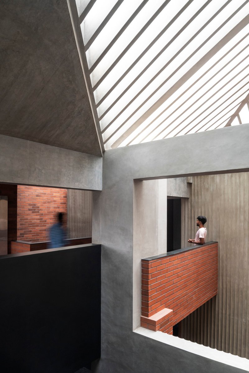 Double-height interior space with slatted skylight casting diagonal shadows across concrete walls and brick balcony where a person stands