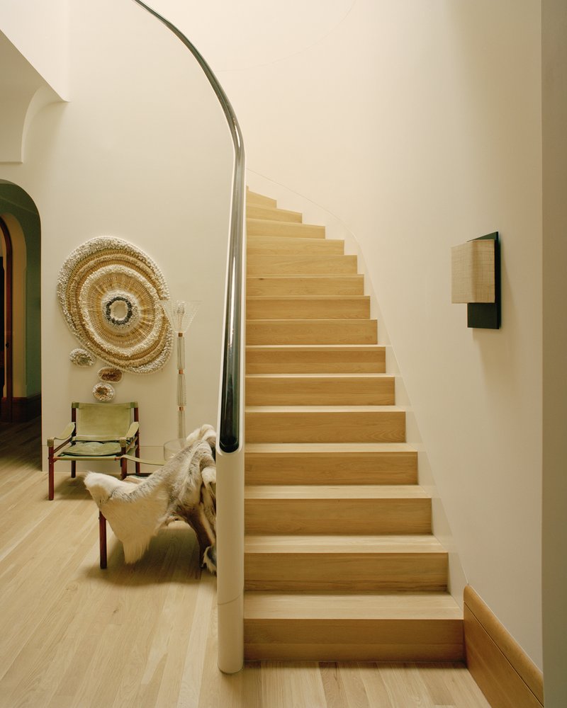 Oak staircase with curved steel handrail ascending beside white walls and a chair with woven wall hanging