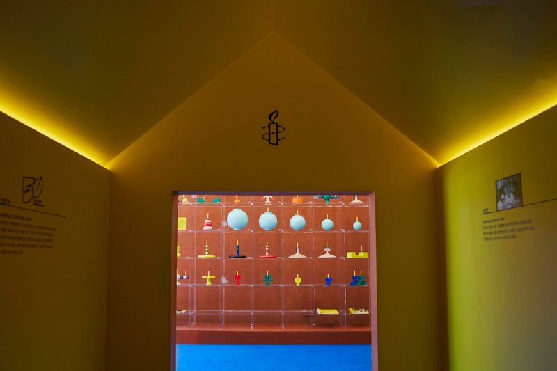 Doorway framing a display vitrine with suspended objects, yellow cove lighting illuminating the gabled ceiling