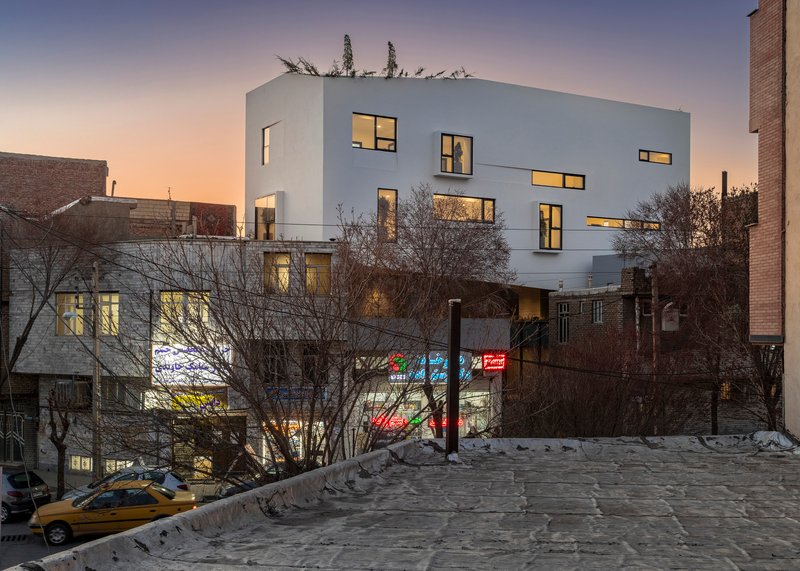 White facade with scattered window openings lit from within at twilight over bare winter trees