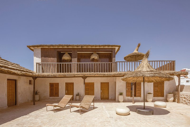Two-story facade with timber shutters and upper balcony overlooking terrace with lounge chairs and thatch umbrellas