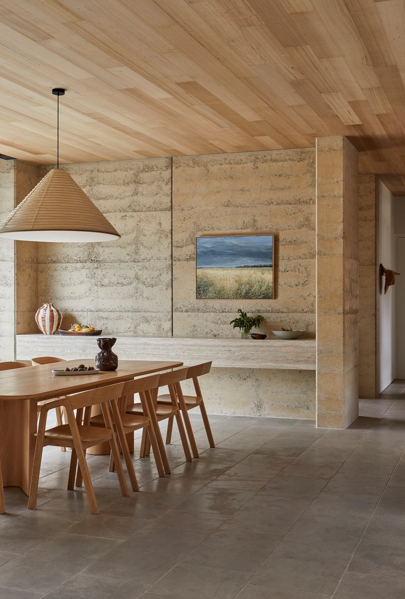Dining area with rammed earth walls, timber plank ceiling and large pendant light above an oval table