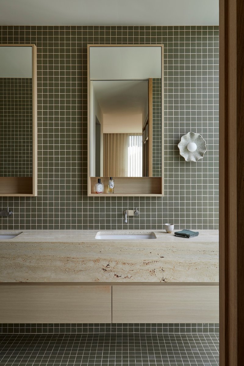 Bathroom vanity with olive green mosaic tile and travertine countertop below framed mirrors