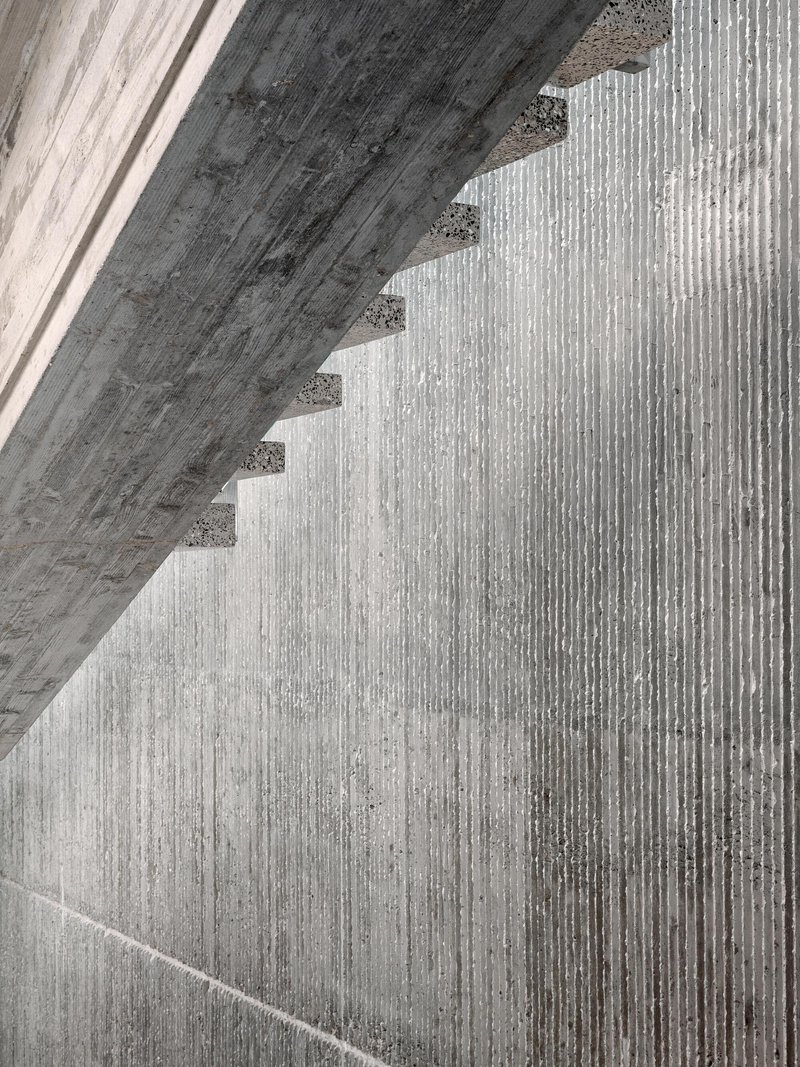 Close-up of board-formed concrete stair soffit casting diagonal shadows across ribbed wall