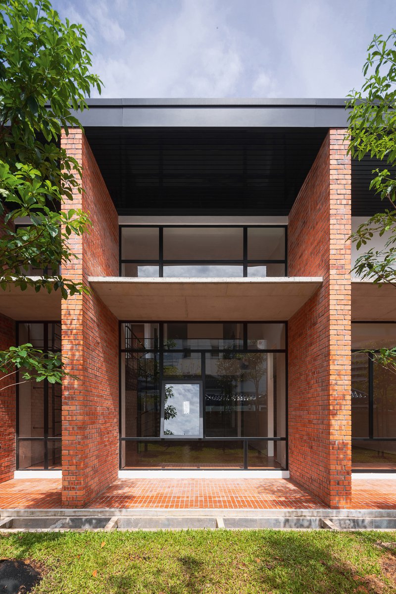 Front facade with red brick columns framing a glazed entrance and recessed upper balcony under lush green trees