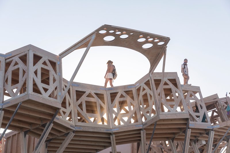 Perforated timber canopy atop geometric wooden framework with visitors silhouetted against the sky