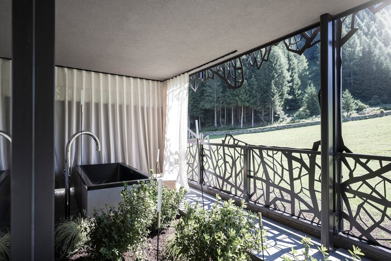 Covered balcony with geometric metal railing, white curtains, and planted greenery facing the forest