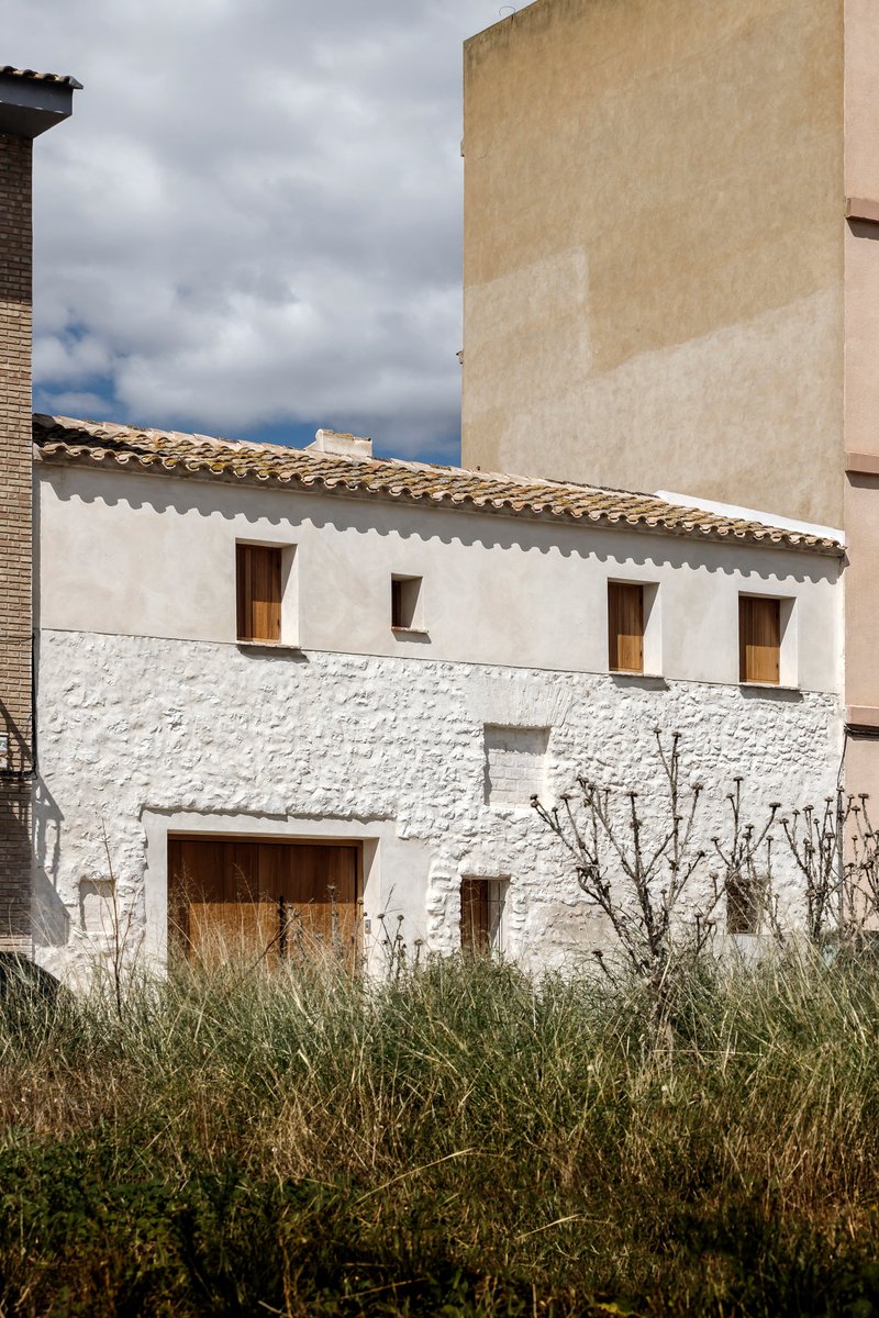 White rendered two-story facade with timber openings and terra-cotta tiled roof under cloudy sky