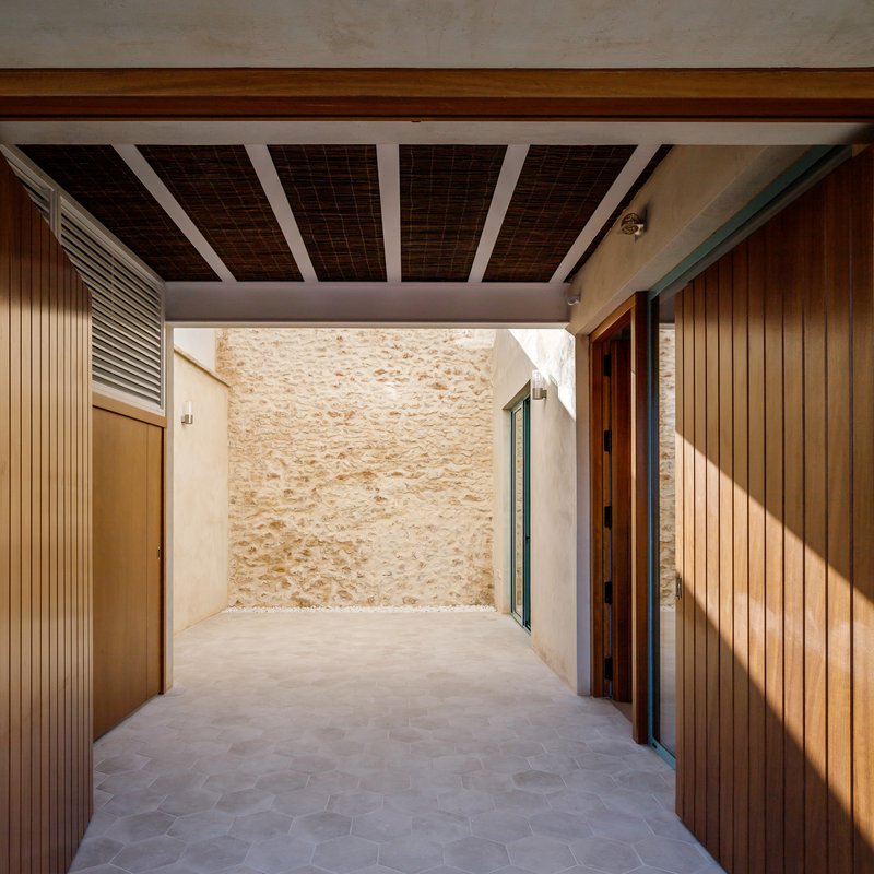 Covered entry passage with timber-slatted ceiling revealing stone courtyard wall in afternoon light
