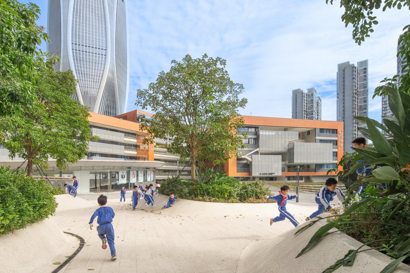 View of the orange-banded school building across a sand play area with children running and trees framing the scene