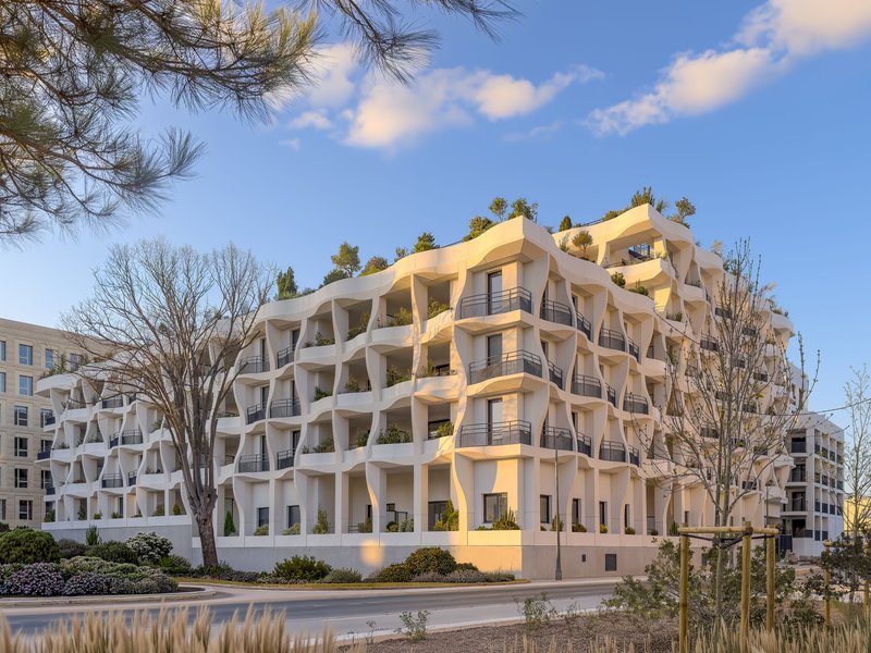 Street view of the undulating white facade with planted balconies framed by bare winter trees