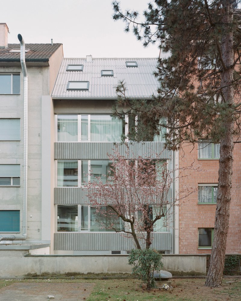 Facade of corrugated metal cladding with tilted dormers framed by a bare tree and an evergreen in winter