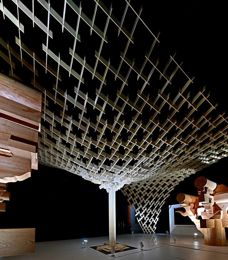 Funnel-shaped timber canopy descending from a diagrid ceiling in a dimly lit gallery