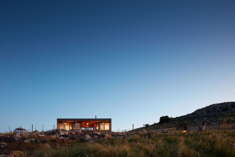 Oblique view across native grasses and limestone boulders toward the glowing structure at dusk