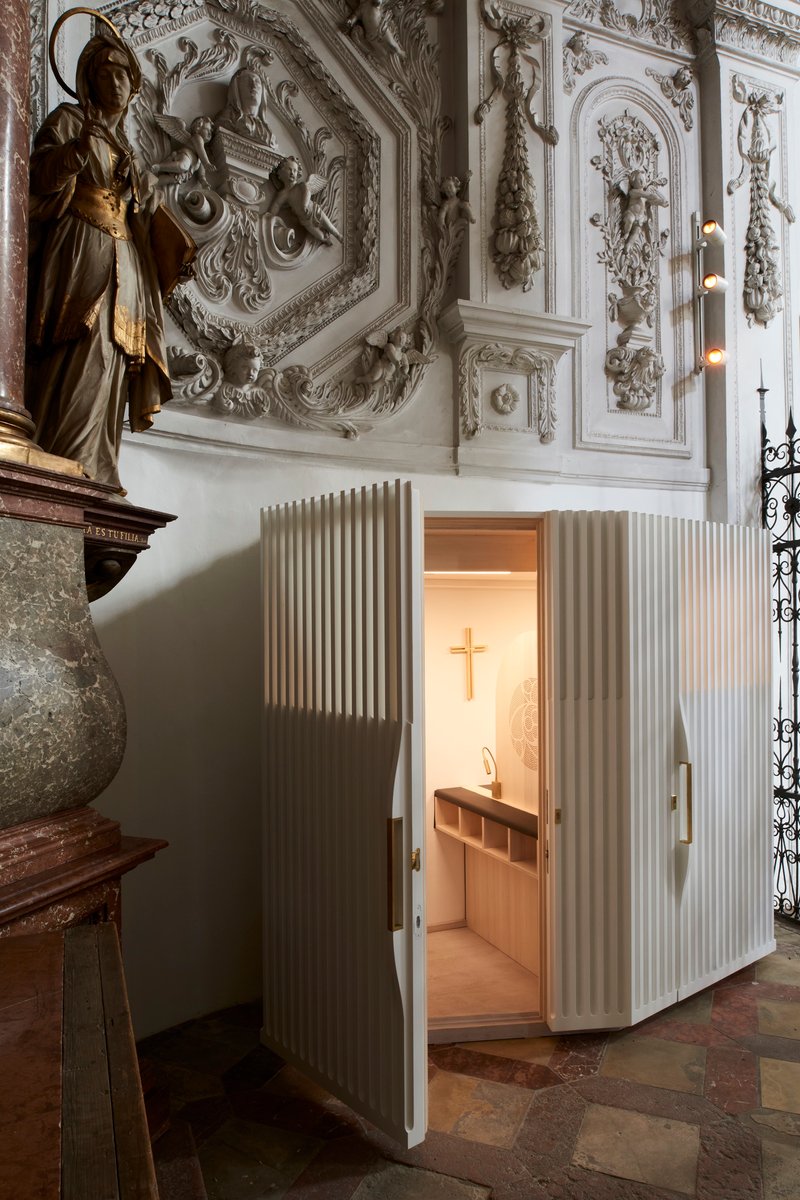 Open doorway in the ribbed timber enclosure revealing the lit confessional interior with cross and seating