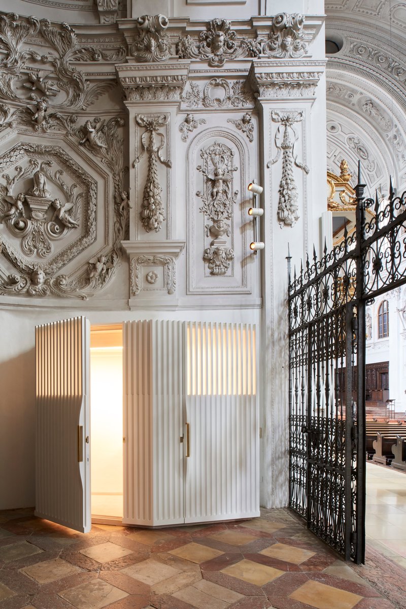 White fluted confessional booth with open door showing illuminated interior and brass handles in the church nave