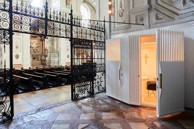 White confessional booth with illuminated cross and vertical fins beside ornate wrought iron gates and pews