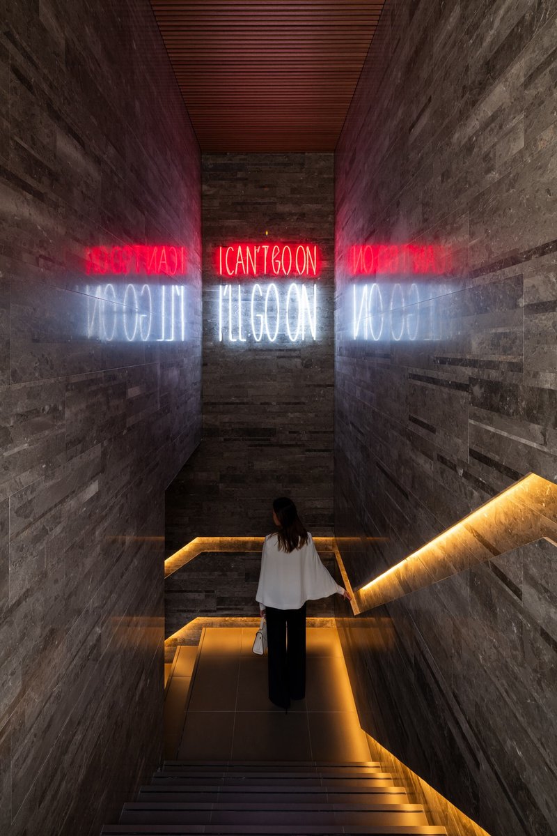 View down a concrete stairwell with red and white neon text reflected on board-formed walls