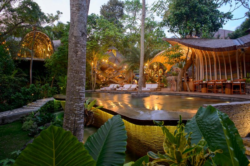 Courtyard pool at dusk framed by palm trunks and curved timber pavilions with uplighting