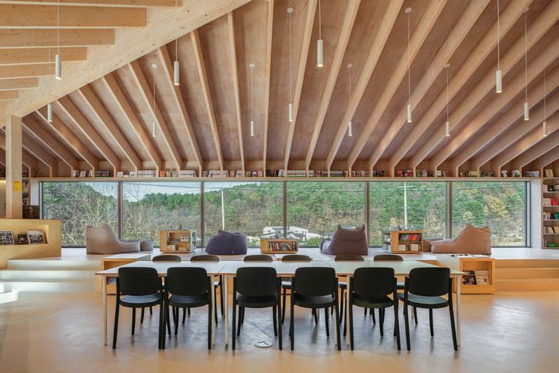 Communal table with black chairs beneath fanned timber rafters and continuous glazing to the hillside