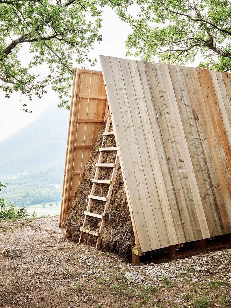 Triangular timber pavilion with vertical wood cladding and straw infill beneath a canopy of spring foliage