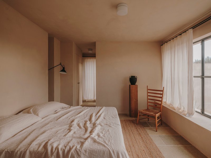 Bedroom with pale plaster walls, sheer curtains and a ladder-back chair beside a timber cabinet