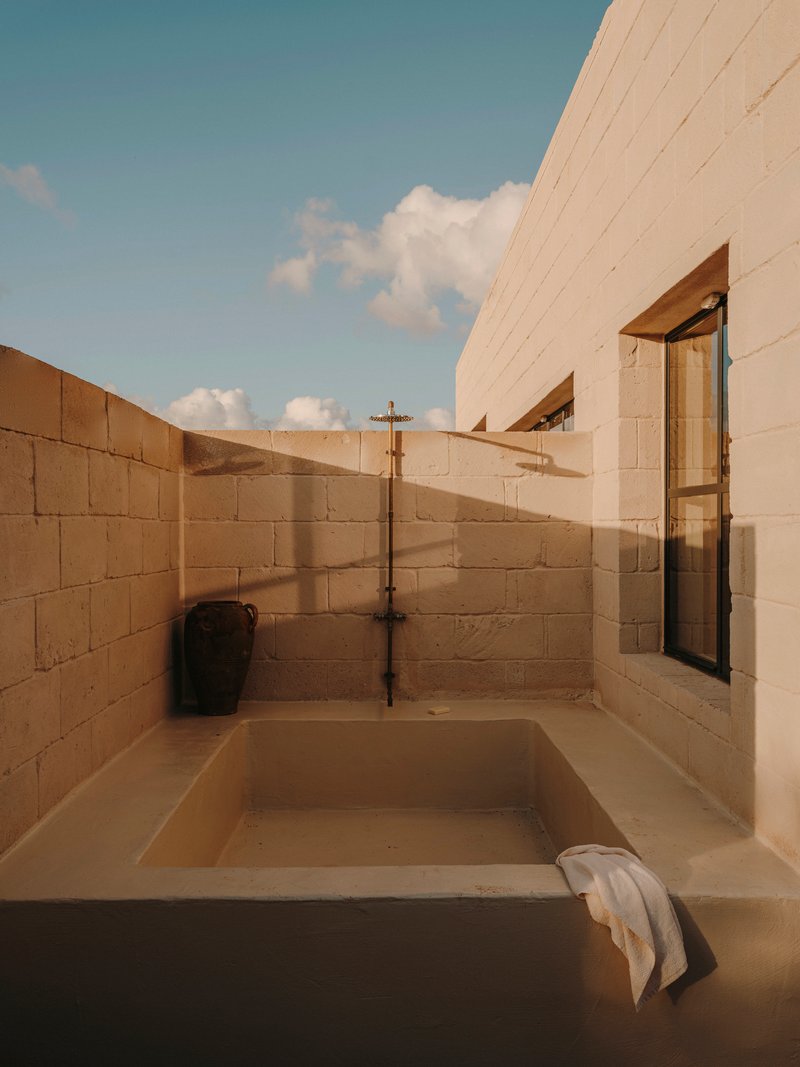 Outdoor sunken bathtub enclosed by concrete block walls with exposed brass plumbing under afternoon sky