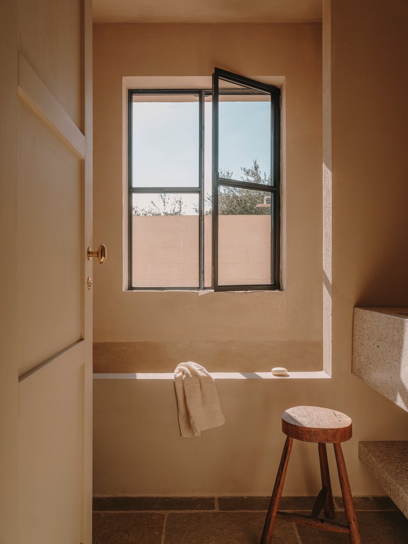 Built-in bathtub beneath a casement window overlooking a terrace with olive trees and wooden stool