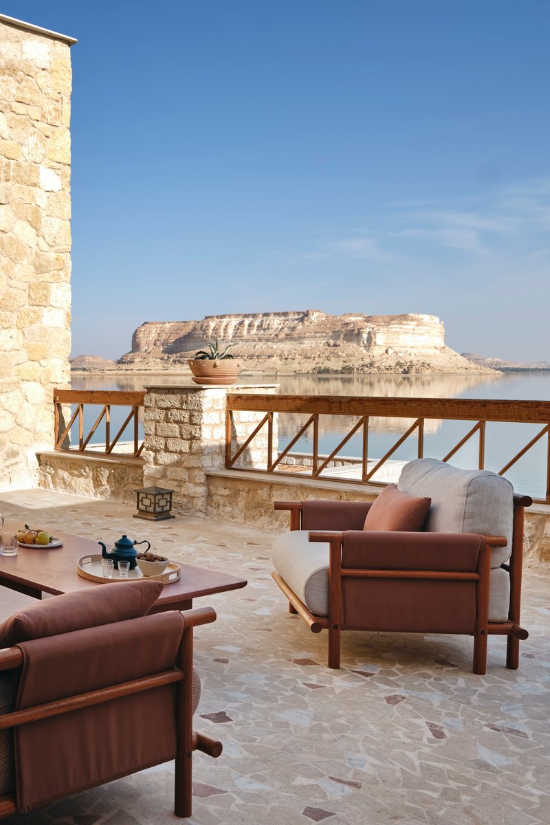 Limestone terrace with timber armchairs overlooking a distant mesa across calm water in bright daylight