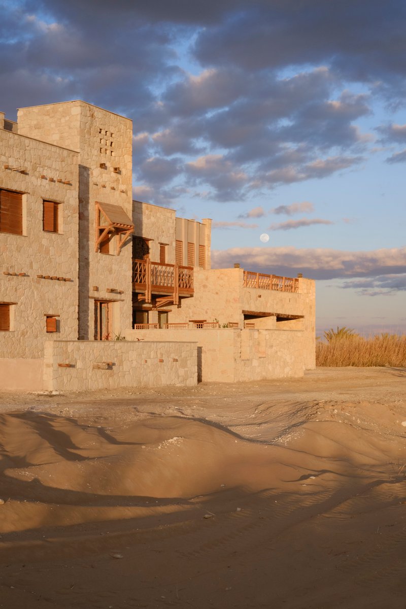 Clustered limestone volumes with timber balconies and openings rising from wet sand under evening clouds