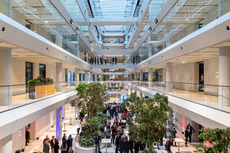 Multi-story atrium with glass-railed balconies encircling a central courtyard planted with trees and filled with visitors