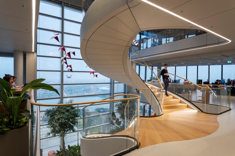 Spiral staircase with white paneled soffit and brass handrail ascending past floor-to-ceiling windows with red paper birds