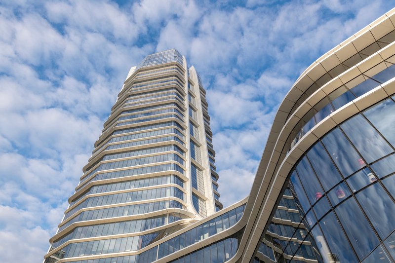 Upward view of the horizontally banded glass tower and curved podium facade under scattered clouds