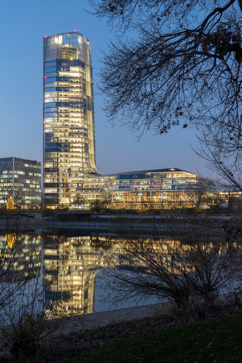 Evening view of the illuminated tower and waterfront podium reflected in the river framed by bare branches